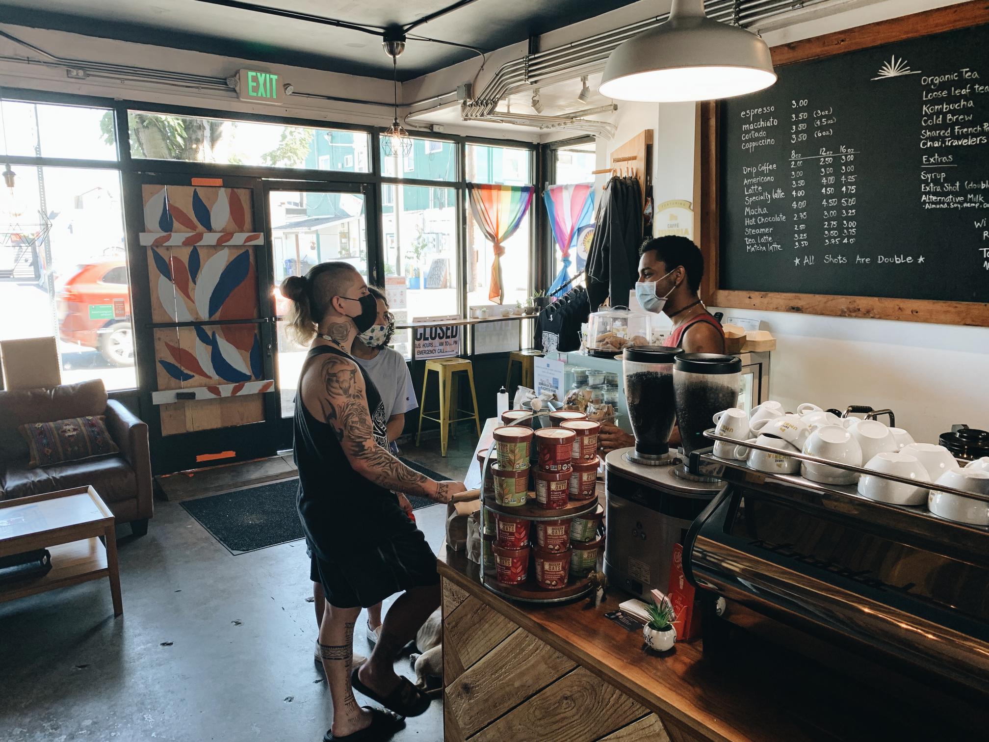 Two community members stand inside a coffee shop in West Seattle in June 2021. They are greeted by an employee standing behind the counter, under a large menu with drinks written on a chalk board. All three people pictured are wearing masks, and the street is visible in the background, through large windows.