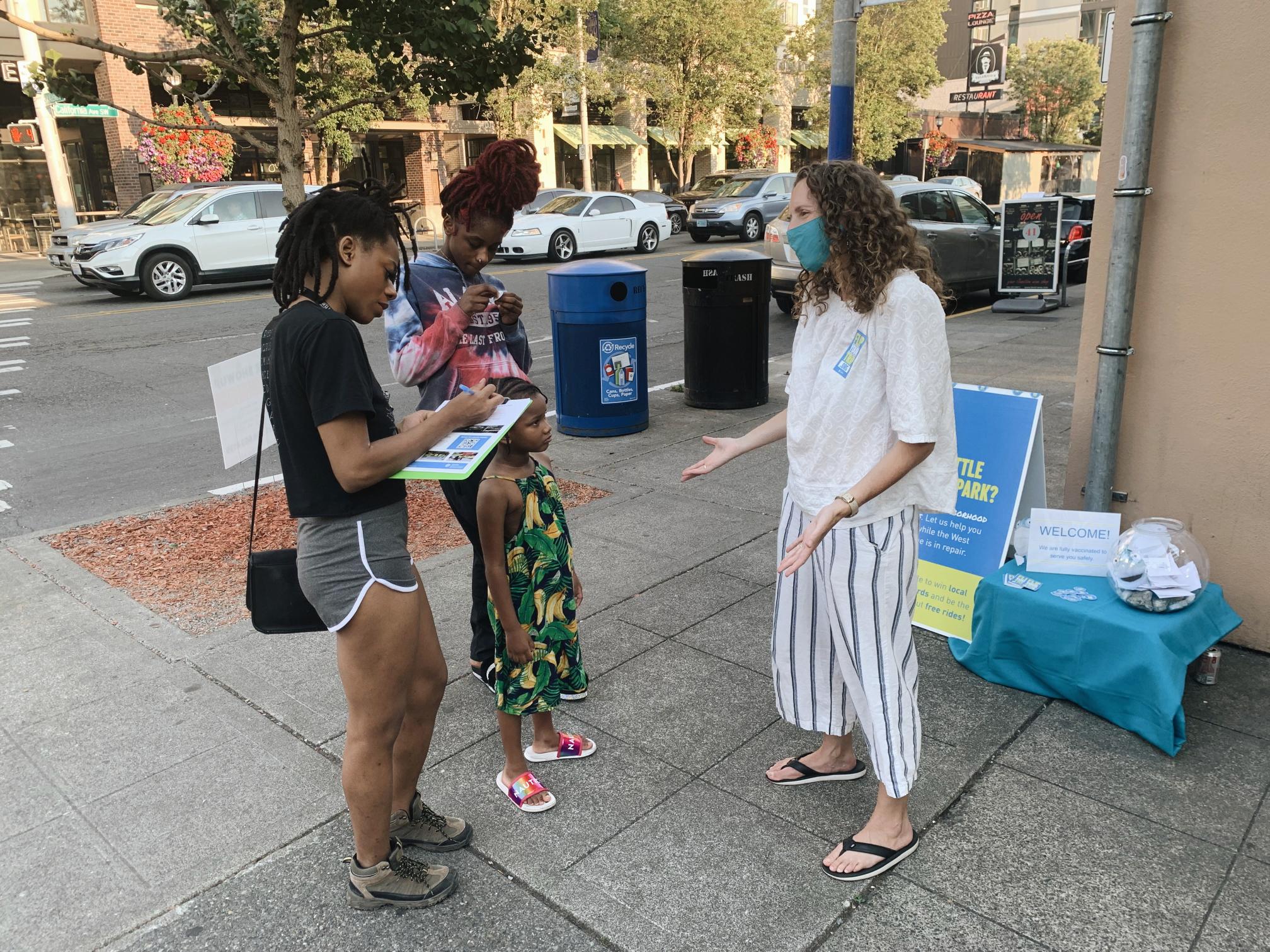 A member of the project team speaks with several community members in West Seattle in June 2021. A women and two younger residents are visible filling out a survey, and speaking with the project representative. Several cars and sections of sidewalk and roadway are visible in the background.