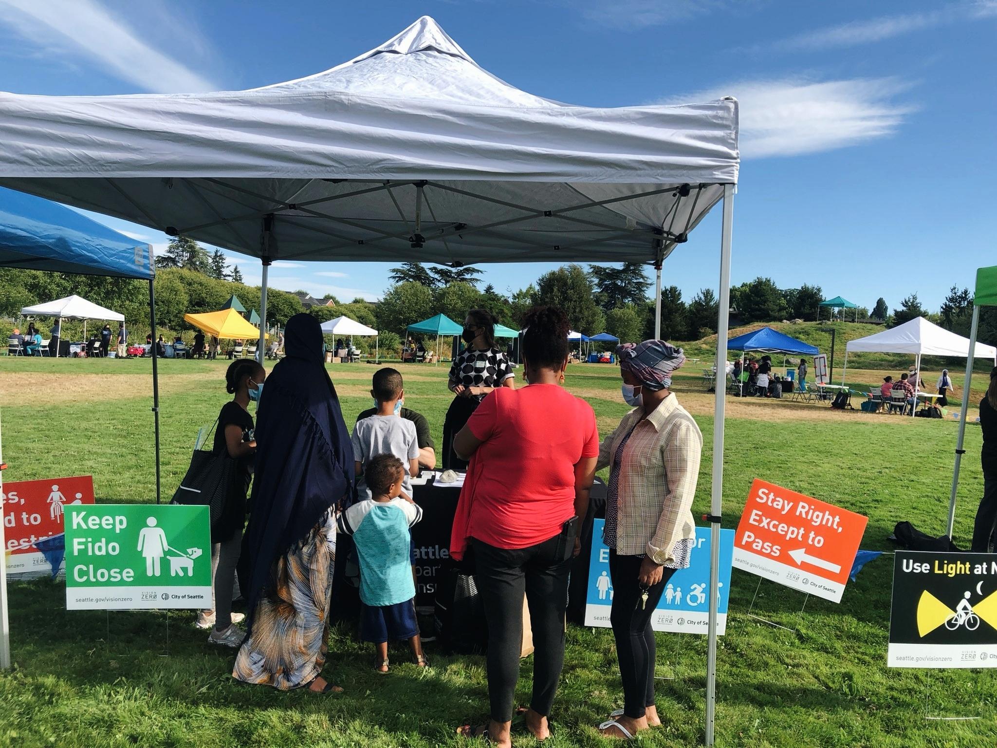 Community members visit an outreach booth in the High Point neighborhood during outreach in summer 2021. Several community members, adults and kids, are visible speaking with the project team on a large grass field, with blue sky and clouds visible in the background. People pictured are gathered under and in front of a white pop-up canopy.