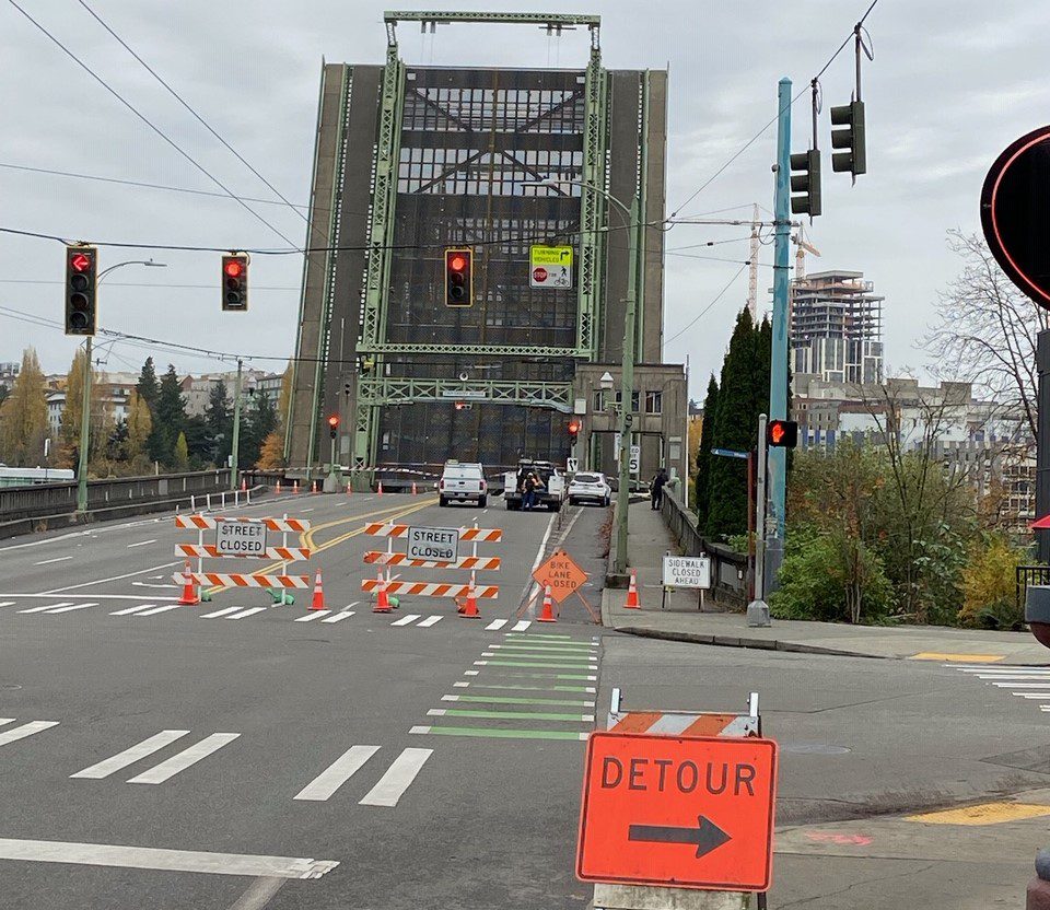 Photo of the University Bridge during the closure, with the bridge deck elevated. Orange detour route signs are visible in the foreground. Vehicles of crews working at the bridge are visible near the bridge, as well as several trees in the background.