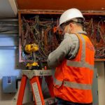 An engineer works on an electrical panel in the University Bridge in Seattle. The worker is wearing an orange safety vest and white hard hat, and eye protection, as he works on the electrical wiring shown in the photo.