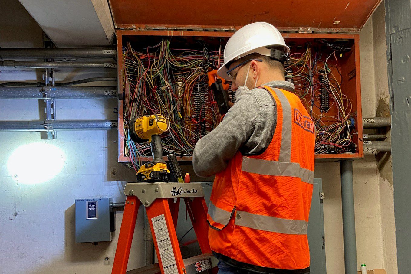 An engineer works on an electrical panel in the University Bridge in Seattle. The worker is wearing an orange safety vest and white hard hat, and eye protection, as he works on the electrical wiring shown in the photo.
