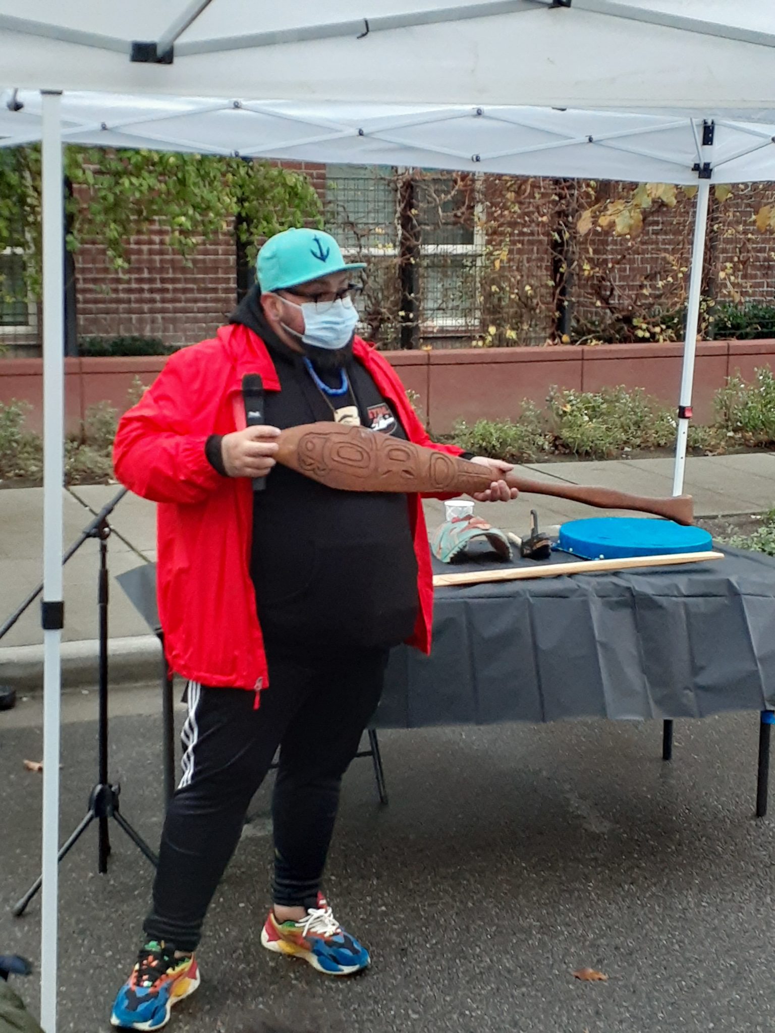 Storyteller Ty Juvinel (Tulalip), shows a traditional paddle that's been in his family for more than 100 years. Ty is wearing a blue mask and holding a microphone while he shares about the paddle. A table and canopy tent are visible in the background behind him.