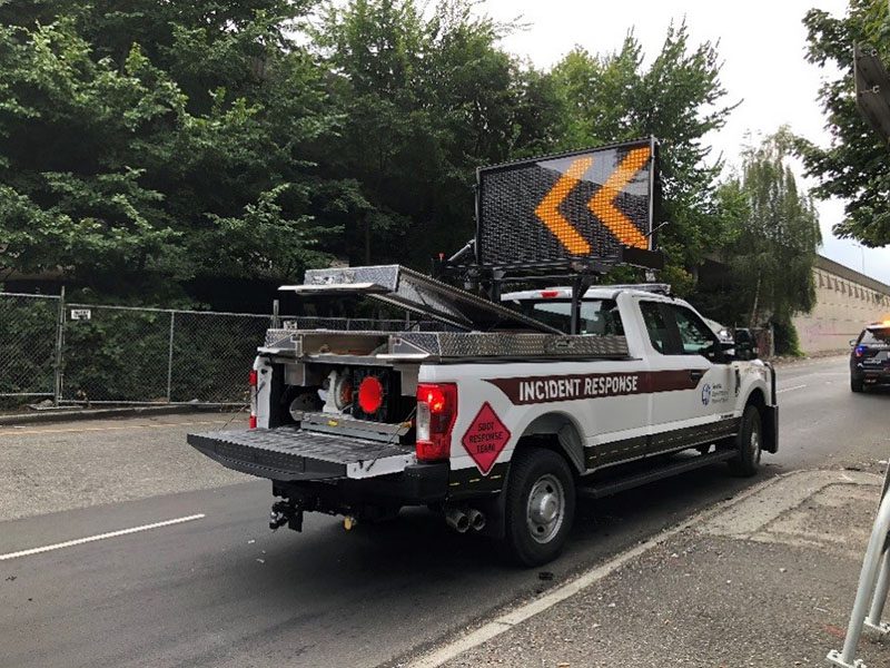 A Seattle Department of Transportation Response Team truck works in the roadway in Seattle. An electronic sign above the vehicle indicates to drivers to merge left, with two large left-direction arrows. The roadway and several large trees are visible in the background.
