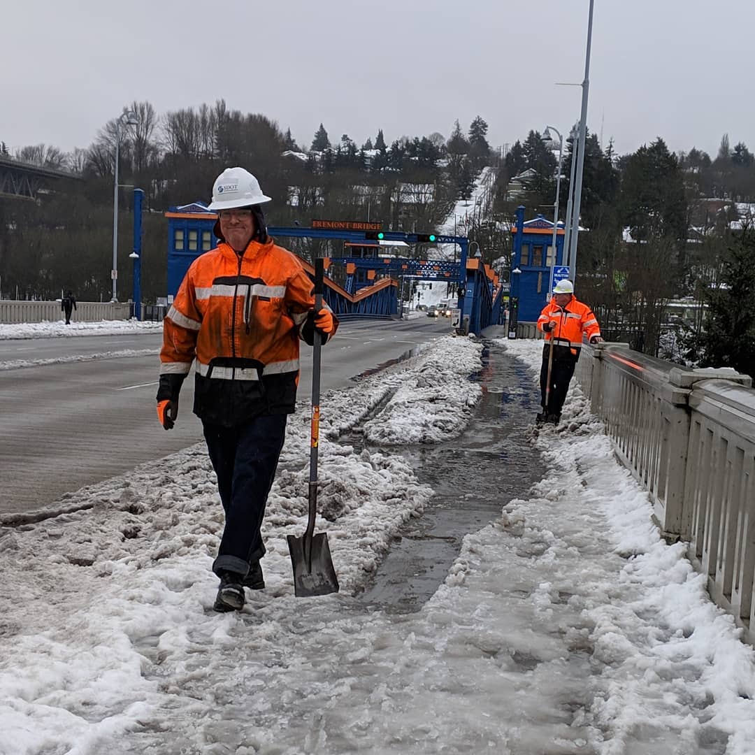 Seattle Department of Transportation workers use shovels to clear a path for people walking, rolling, and biking across the Fremont Bridge in Seattle during the February 2019 snow storm. The two workers wear bright orange safety coats and white hard hats while they work.