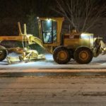 A Seattle Department of Transportation worker operates a grader, which is used to plow snow from city streets. The large piece of equipment is yellow, and travels down a large city street.