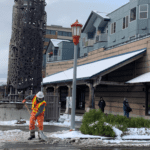A worker wearing an orange safety vest and hard hat works to clear snow from the roadway and a crosswalk along 6th Ave S in Seattle's Chinatown International District. Large buildings are visible in the background, as well as a lamp post.
