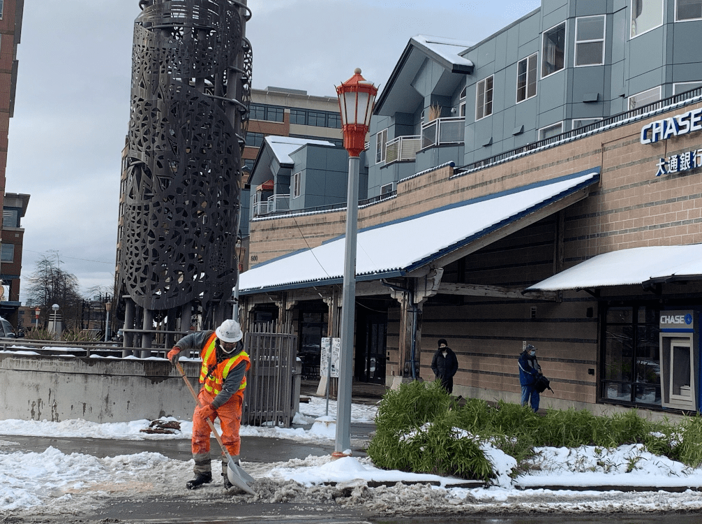 A worker wearing an orange safety vest and hard hat works to clear snow from the roadway and a crosswalk along 6th Ave S in Seattle's Chinatown International District. Large buildings are visible in the background, as well as a lamp post.