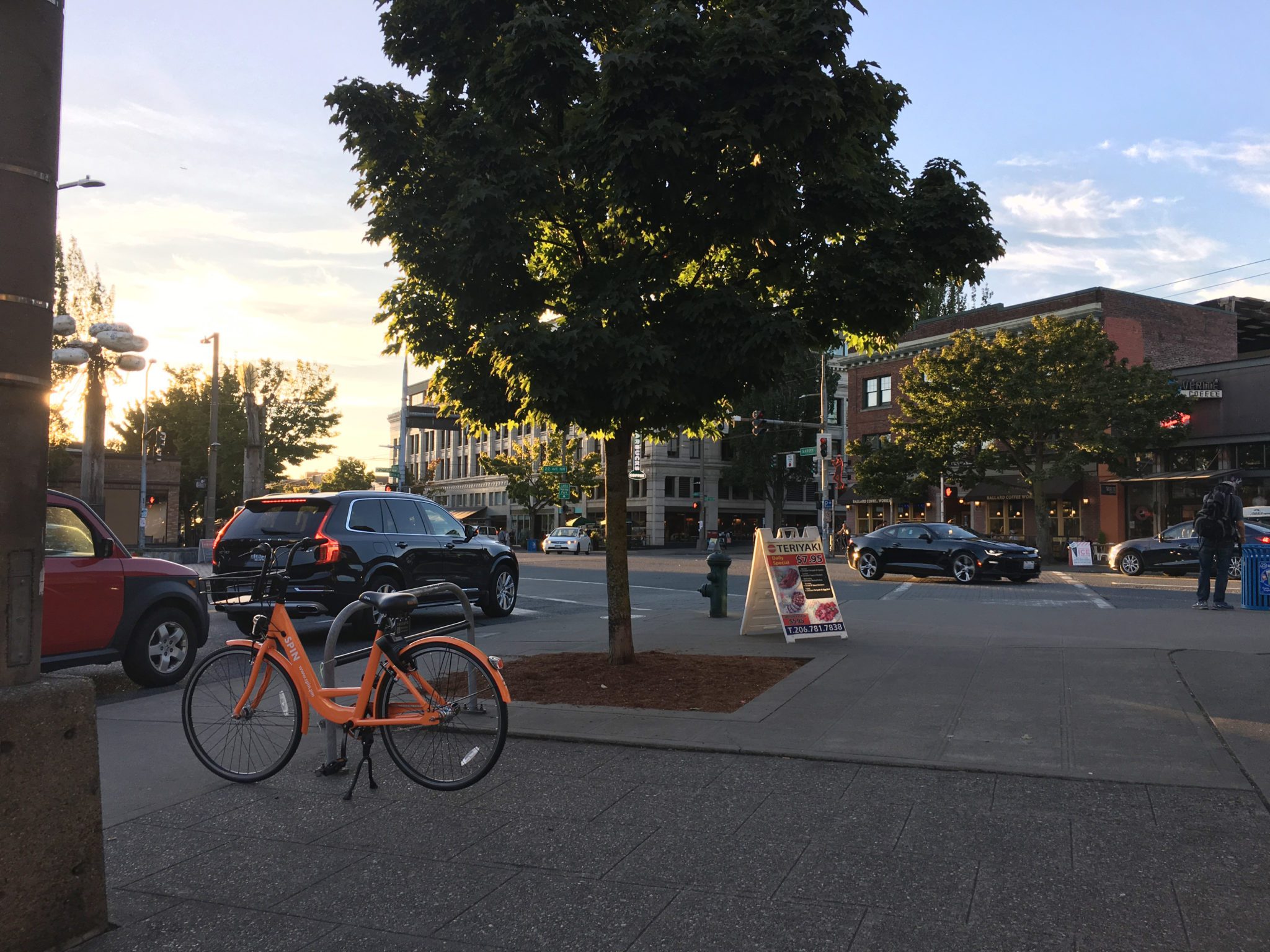 An orange bike share bicycle is properly parked on the sidewalk in Seattle's Ballard neighborhood. The bicycle is visible in the lower left corner of the image. Several buildings, cars, and a large tree are visible throughout the other parts of the image. The photo was taken in the evening.