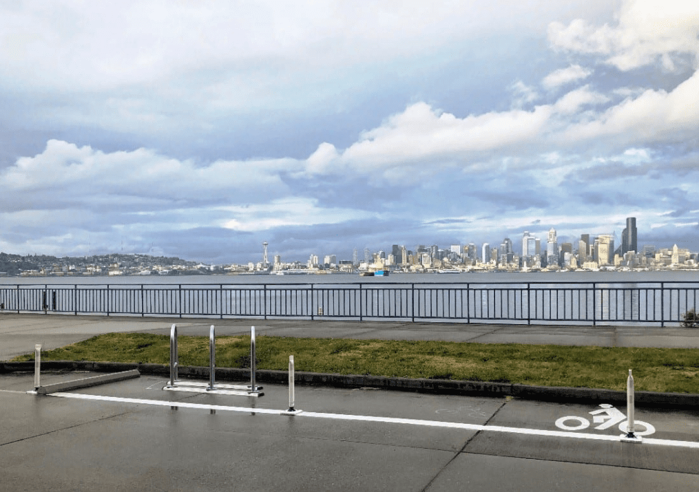 View of a bike parking area and bike parking stall along Alki Ave SW, a main street that runs near the water of Elliott Bay and Puget Sound in West Seattle. Downtown Seattle skyscrapers, the Space Needle are visible in the background, with clouds visible above.