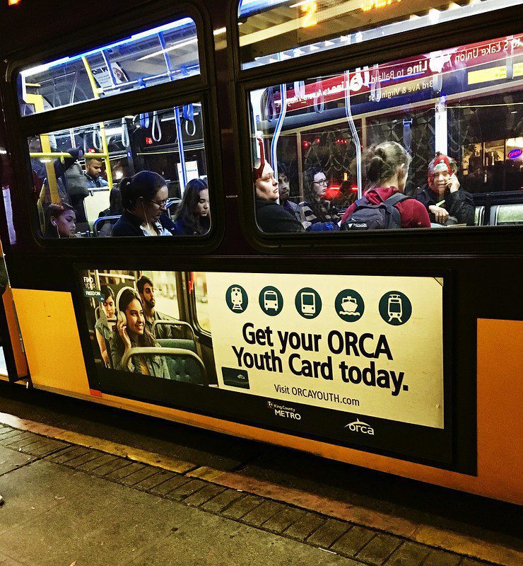 Image of a King County Metro transit bus. Passengers are visible sitting on the bus, while a display sign on the side of the bus advertises an ORCA transit card for youth.