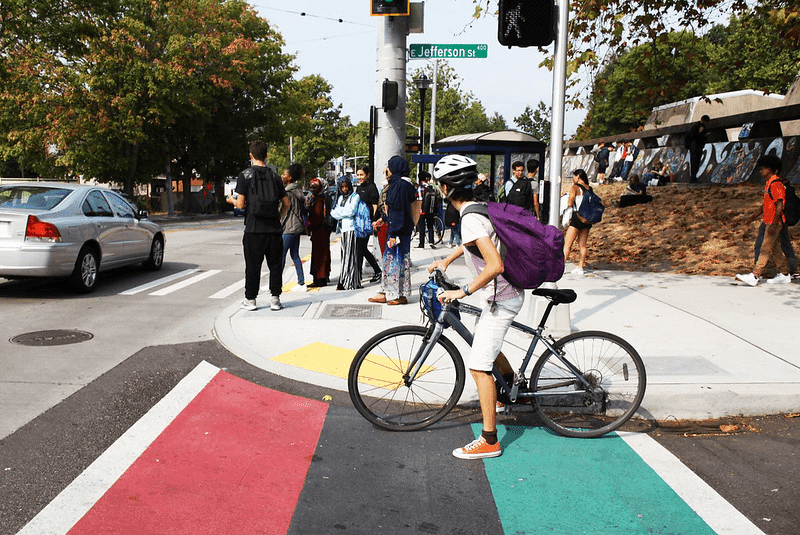 A person bicycling and students walking and standing at a bus stop in Seattle's Central District. A silver car is visible on the left side of the image.