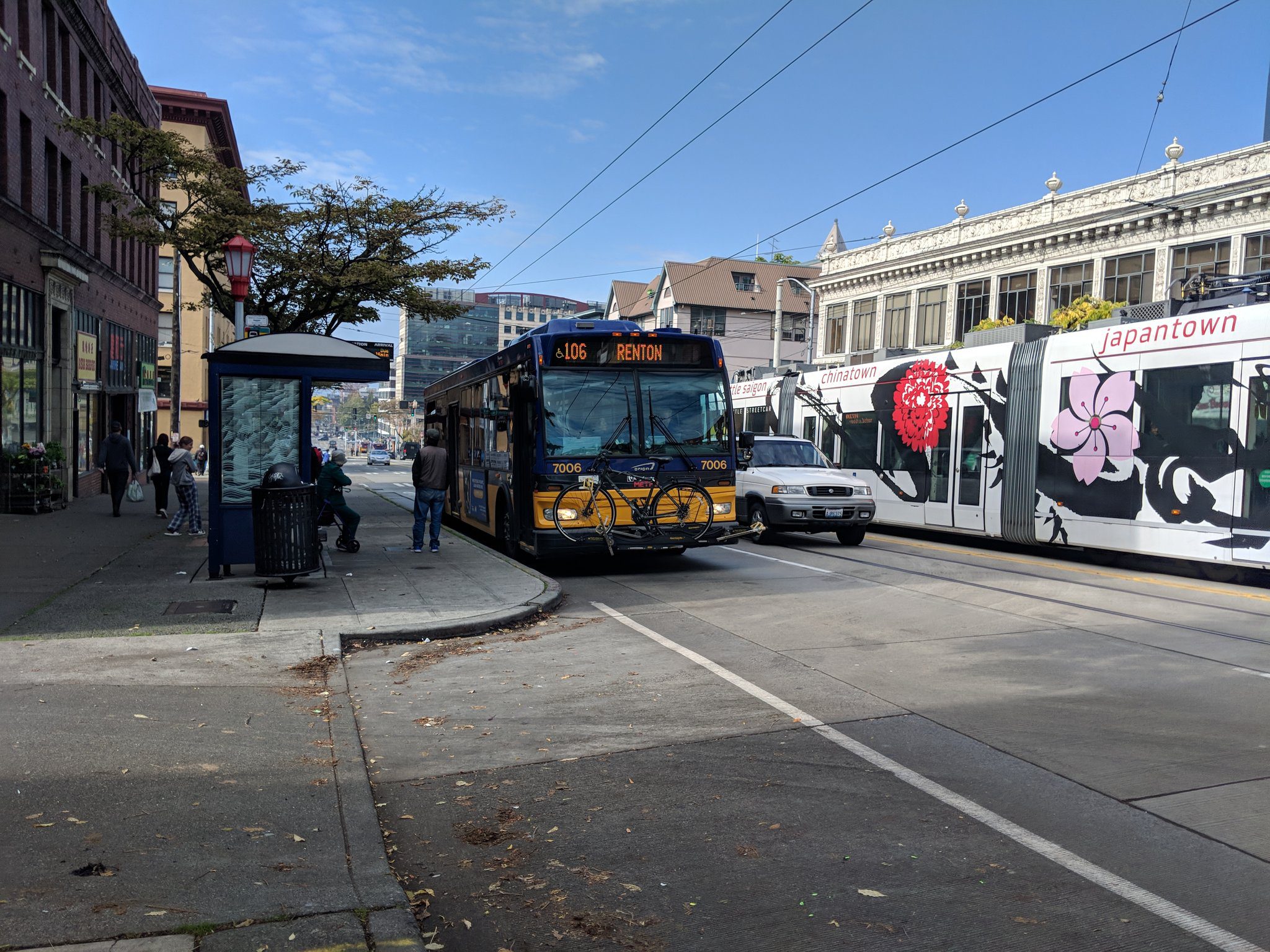 Photo of a King County Metro bus traveling through Seattle's Chinatown International District. The bus is visible in the center of the photo, along with a white vehicle in the middle lane, and the Seattle Streetcar traveling in the opposite direction. Buildings, a bus stop, a tree, and blue sky are visible in the background.