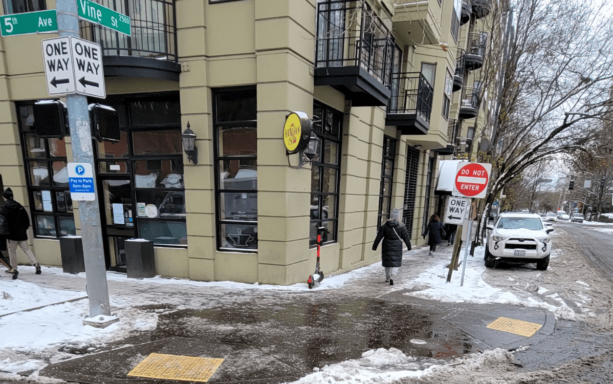 People walk along the sidewalk in Seattle's Belltown neighborhood. Snow and slush are visible in areas of the street and sidewalks, and two sidewalk curb ramps are visible near the botton of the photo. A large building and several trees are also visible.