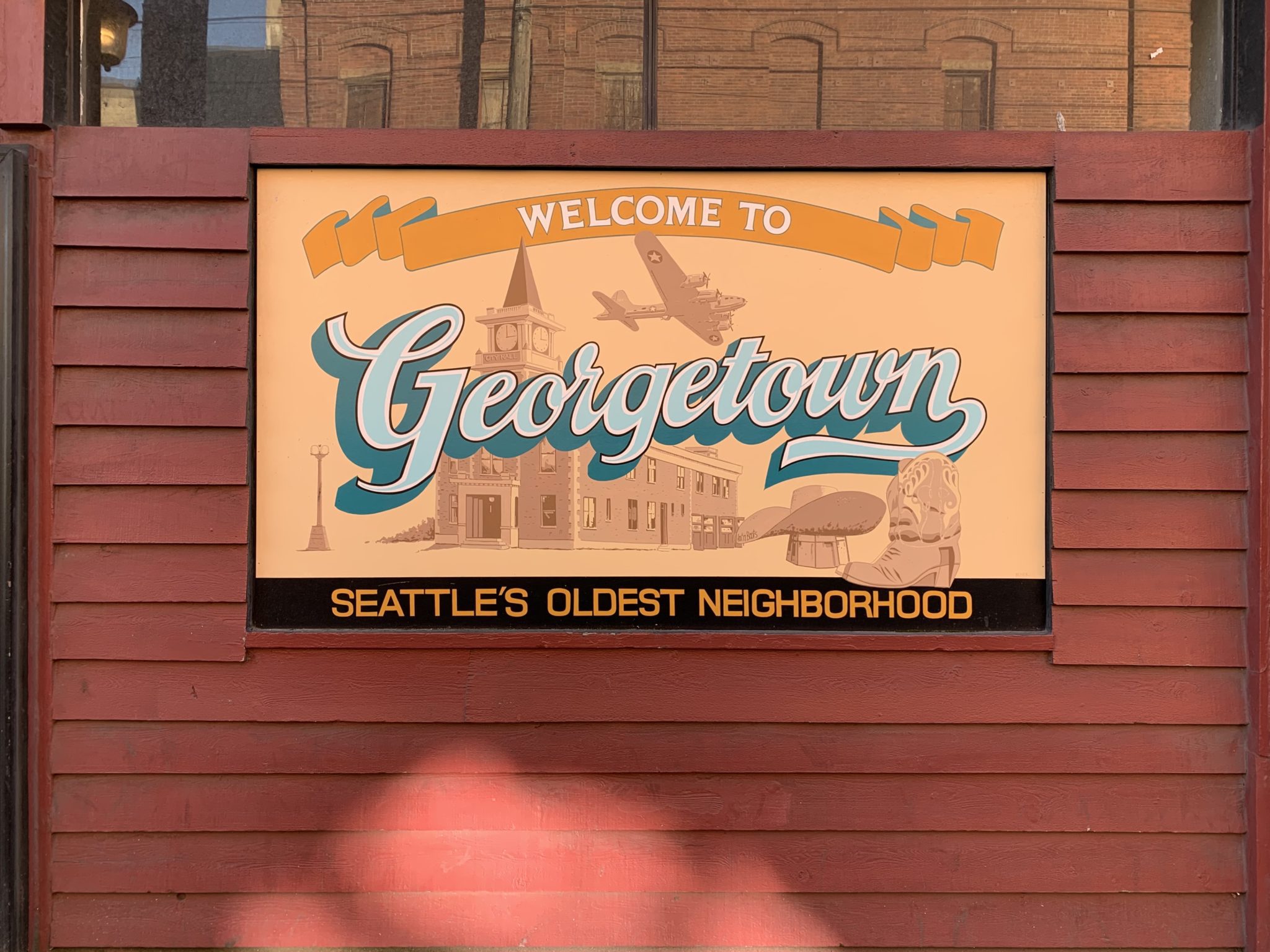A sign board with 'Georgetown' on it in large light blue letters indicates the name of the neighborhood in Seattle. It is hanging on a large red wall, and the sign board has a light orange background.