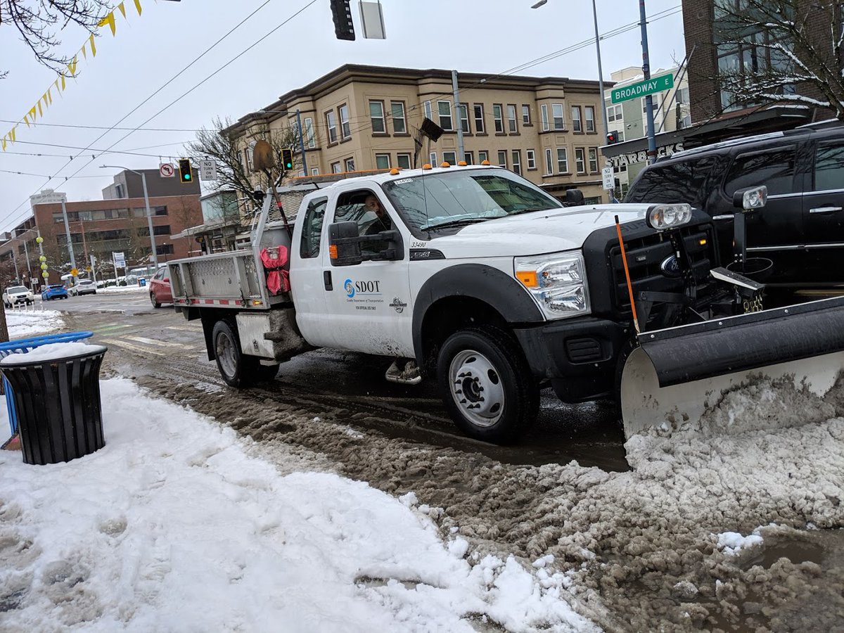 A snow plow truck travels along Broadway E, a major arterial in Seattle. The large white truck is pushing snow to help clear the roadway. Snow is visible in the foreground, as well as several large buildings in the background.
