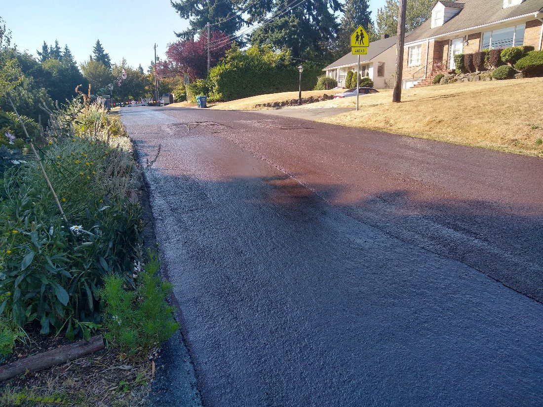 A residential street in Seattle after Slurry Seal treatment was completed. Trees and vegetation are visible on the left and right, and a house is visible in the upper right part of the image.