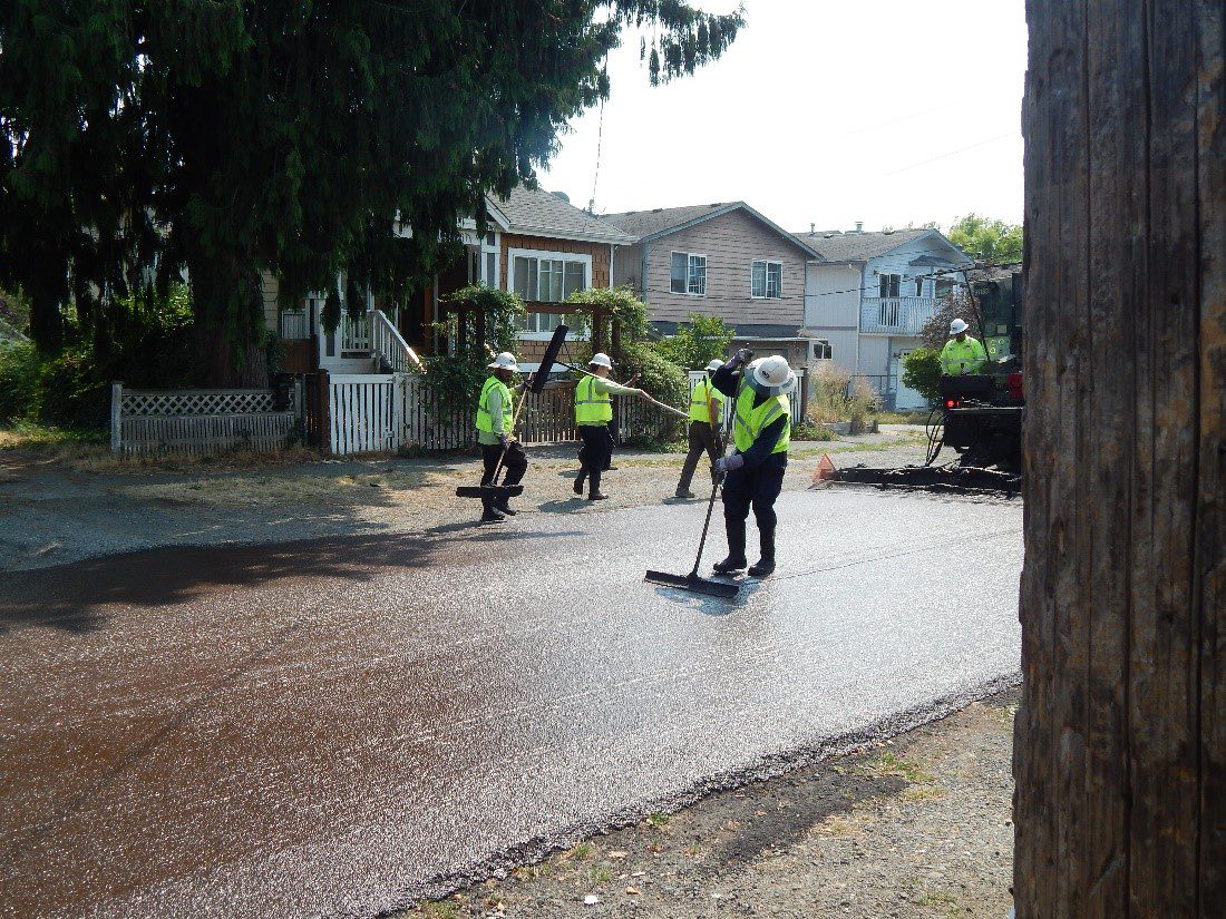 Several workers are visible installing the slurry seal treatment on a local street in Seattle. Five people are visible, all wearing bright yellow safety vests an white hardhats. Homes are visible in the background, as well as tree branches. A pole is visible on the far right side of the image.