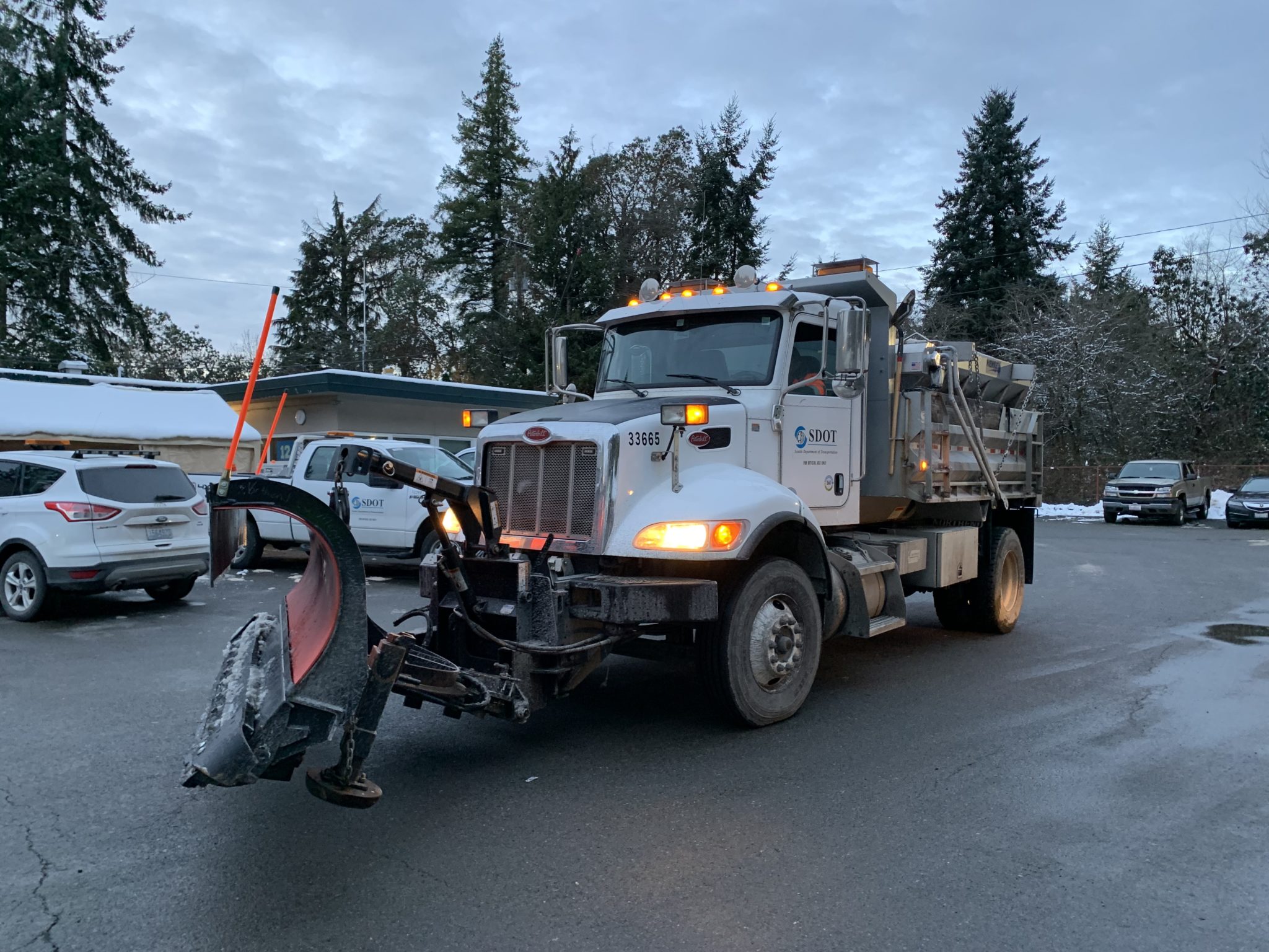 A large snow plow is parked on pavement, ready to return to work clearing streets in Seattle. Several other smaller vehicles and large trees are visible in the background.
