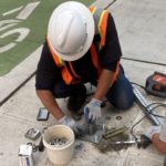 A crew member works to install magnetic and light sensors to detect when a vehicle occupies a curbside space. Our crews installed this equipment in Seattle’s Belltown neighborhood in April 2021. The crew member is wearing an orange safety vest, gloves, and a white hardhat.