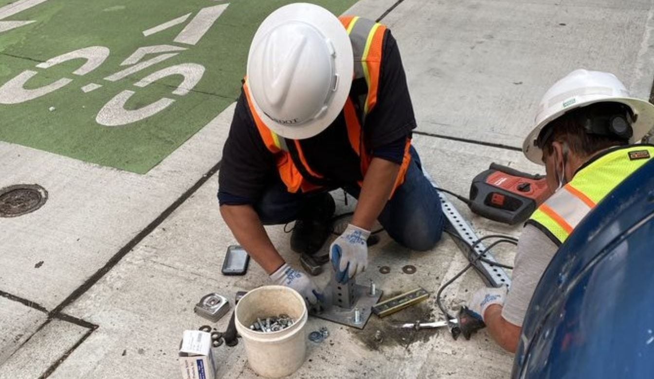 A crew member works to install magnetic and light sensors to detect when a vehicle occupies a curbside space. Our crews installed this equipment in Seattle’s Belltown neighborhood in April 2021. The crew member is wearing an orange safety vest, gloves, and a white hardhat.