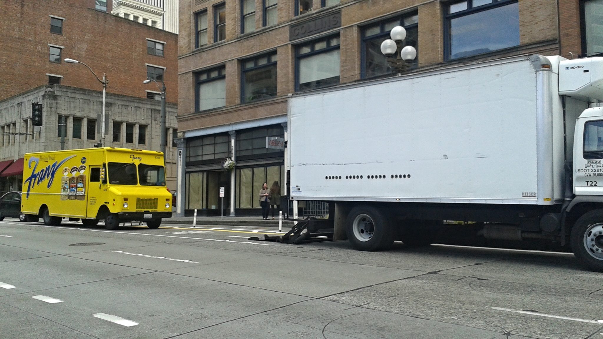 Two trucks parked to deliver bread and other supplies to local businesses in Seattle’s Pioneer Square neighborhood. The truck on the left is yellow, and the truck on the right is white. Large buildings are visible in the background, with the street visible in the foreground.