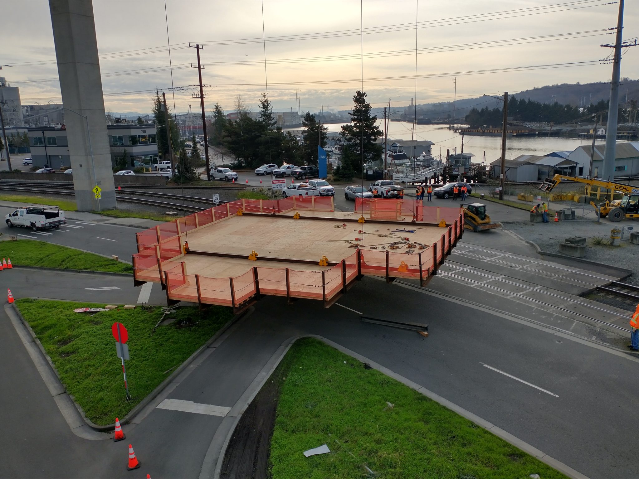 The work platform begins to lift off. Grass, roadway, tracks, and the Duwamish Waterway are visible in the background.