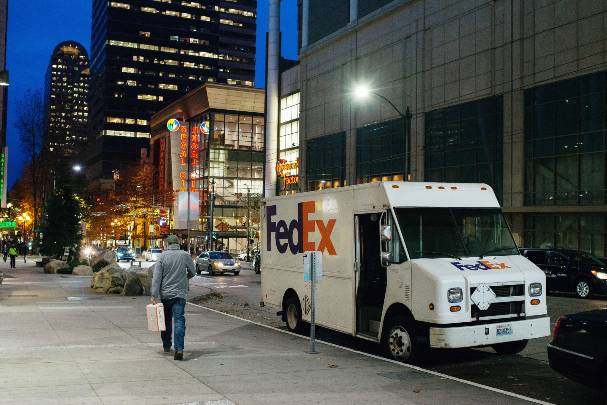 A white FedEx delivery truck is parked in a load/unload zone in downtown Seattle. Large buildings are visible in the background in the evening, and a man walks down the street carrying a bag.