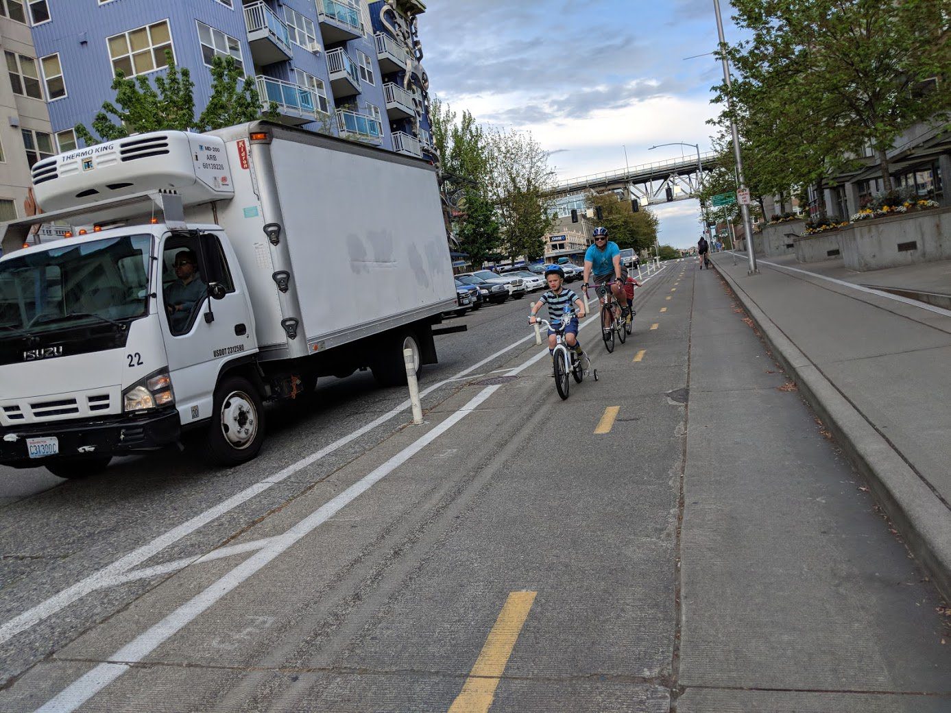 Photo of a family riding their bicycles down a protected bike lane in Seattle. A white delivery truck is visible to the left, with sidewalk and buildings visible to the right.