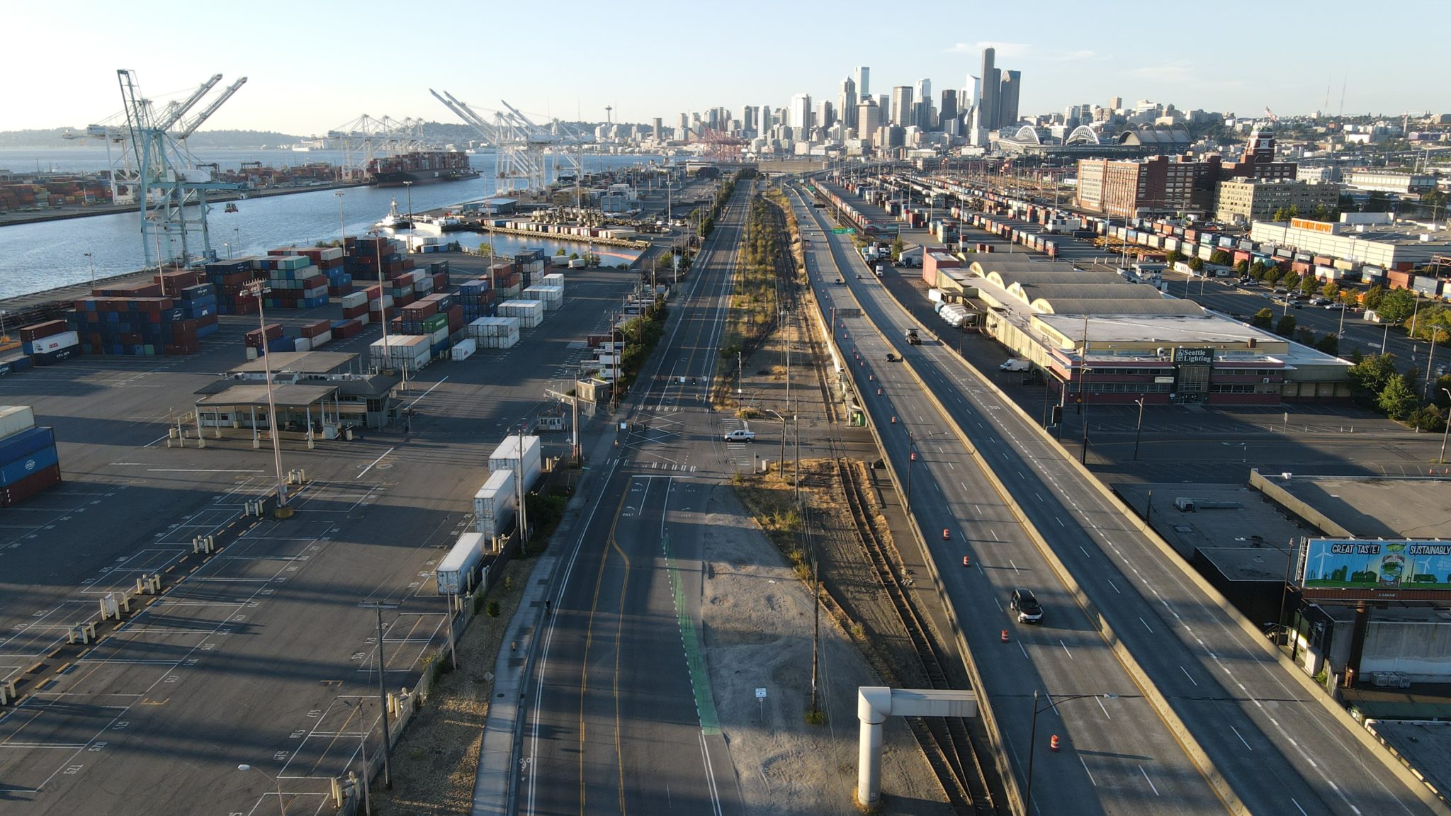 View of East Marginal Way S, looking north toward downtown Seattle. The roadway is visible in the center of the picture, with Port of Seattle cranes visible in the upper left, and downtown Seattle skyscrapers in the background.