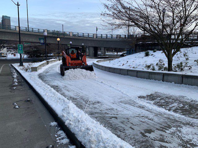 A worker drives a grader, which is a type of snowplow, to help clear a pedestrian walkway near the stadiums in Seattle, on a cold day during recent winter weather. A sidewalk is visible to the left, and a large tree to the upper right.