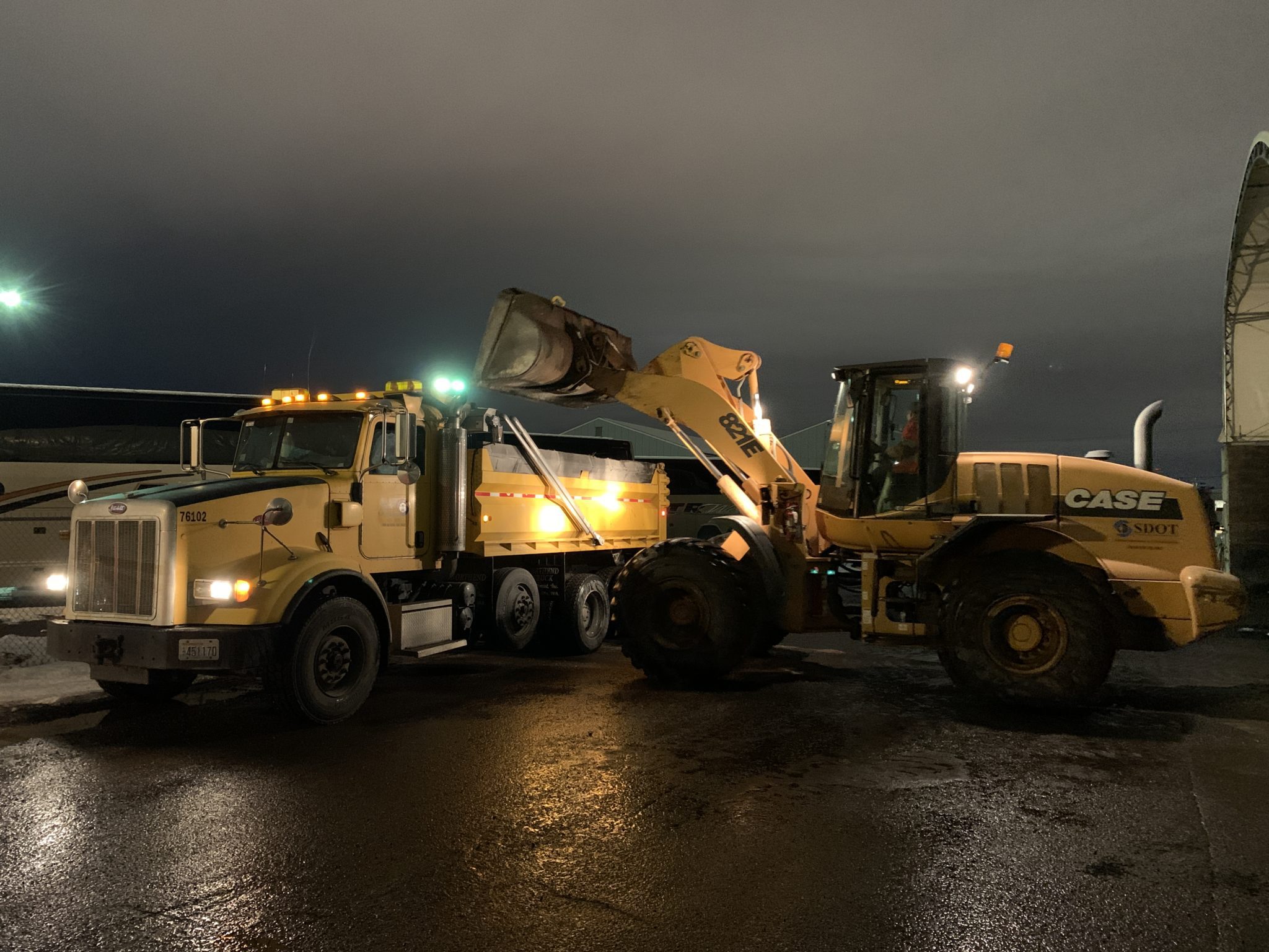 Workers operate two large pieces of heavy machinery to transfer salt to help de-ice streets and other locations in Seattle during the recent winter weather. The photo was taken at night and each vehicle is yellow, with wet pavement visible in the foreground.