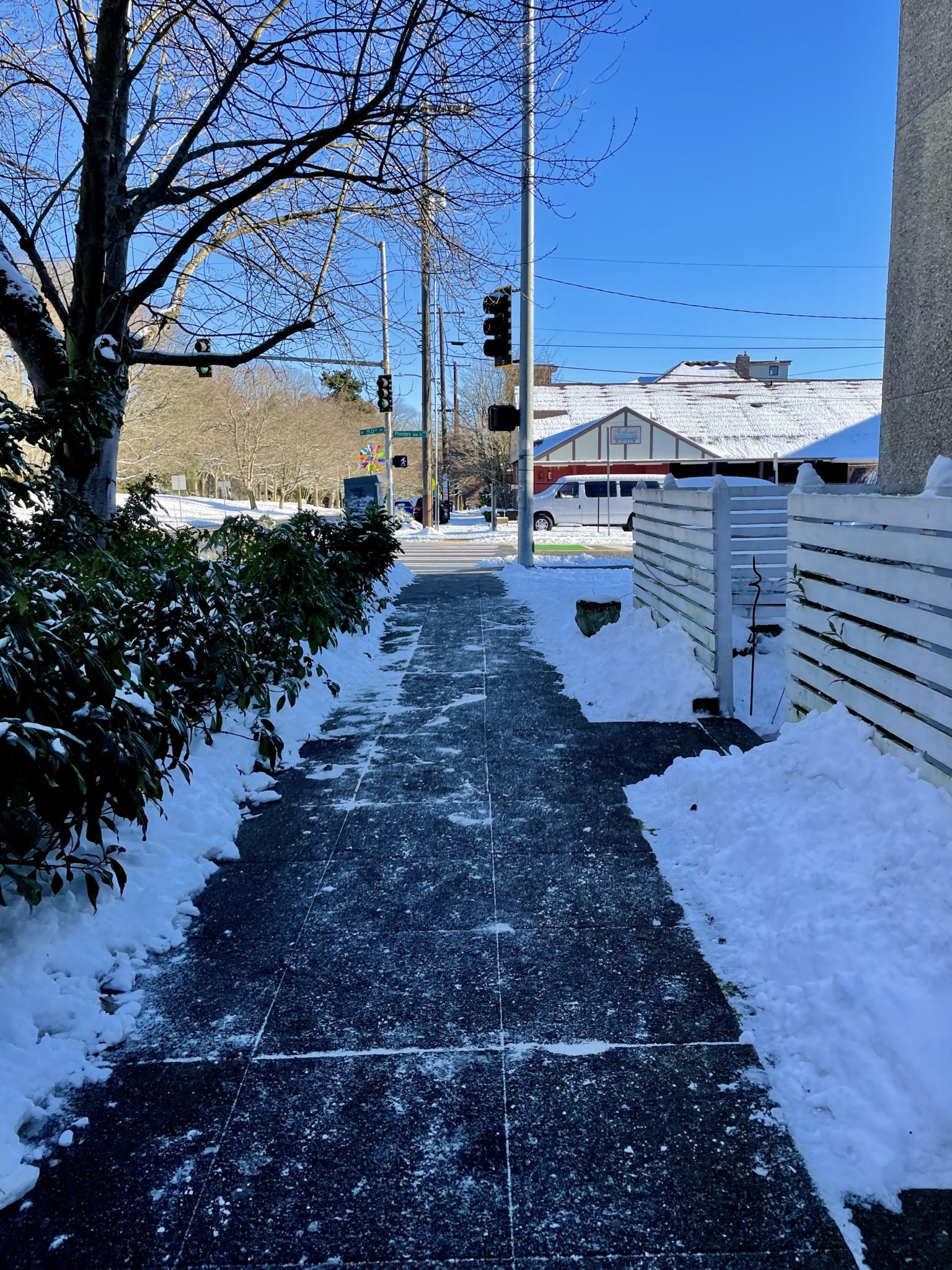 This image shows a sidewalk in north Seattle that had recently been shoveled, creating a clear path for people walking, rolling, and biking in the area. Snow is visible on the ground nearby, as well as clear, blue skies above.