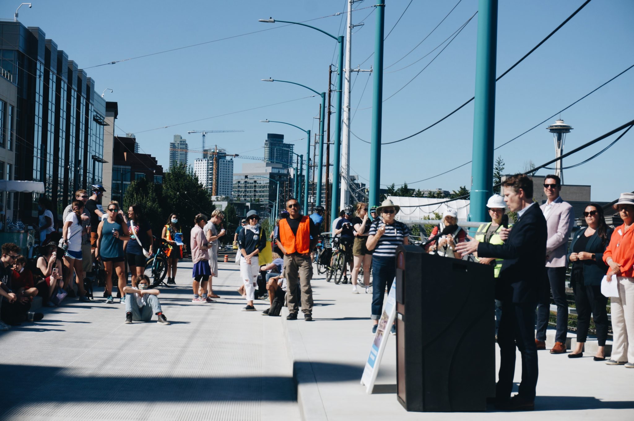 Photo of the grand opening celebration of the new Fairview Ave N bridge in Seattle, in July 2021. Seattle City Councilmember Alex Pedersen is visible speaking at a podium, and neighbors listen to the remarks. The Space Needle is visible in the background.