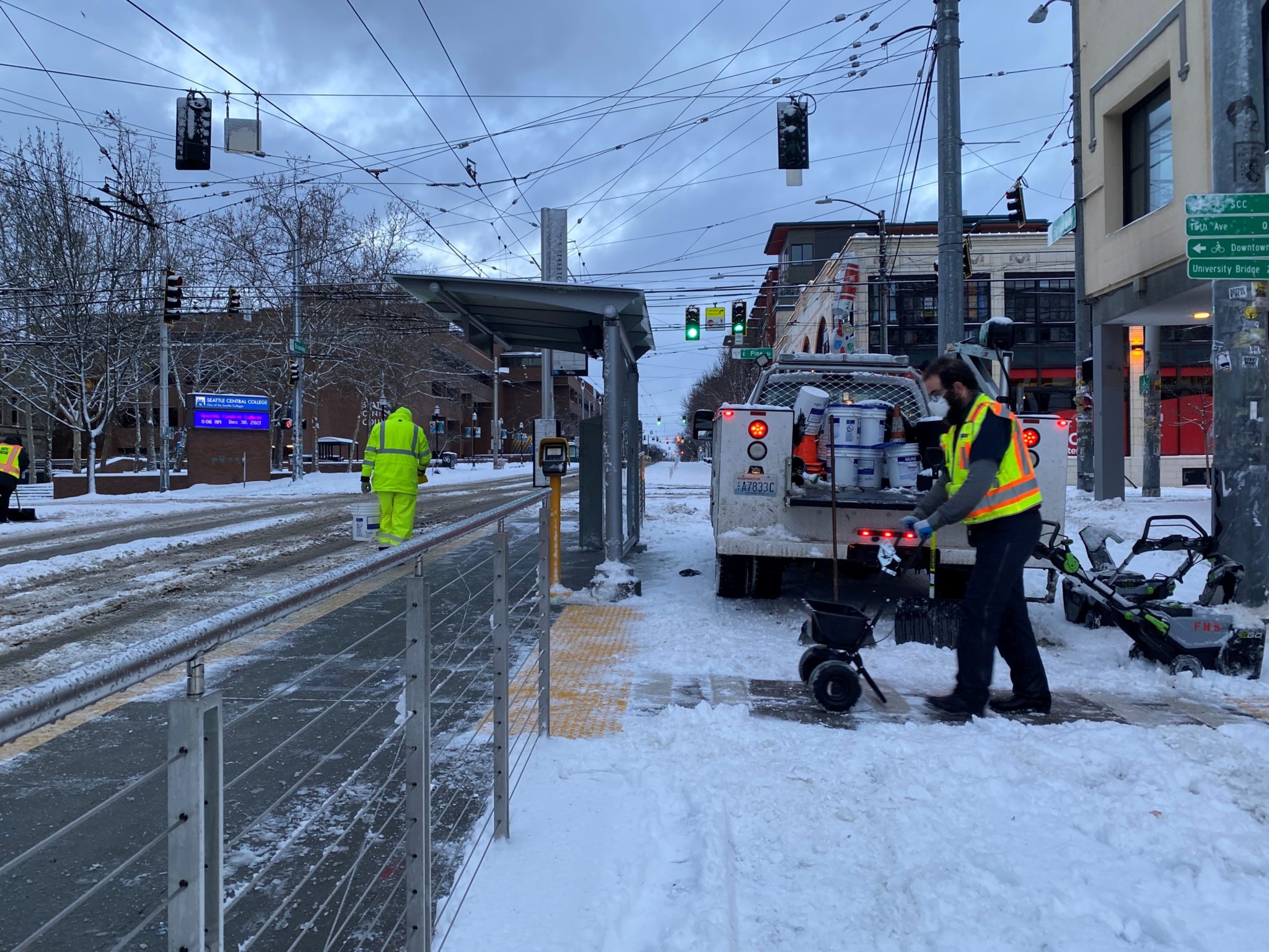 Two workers clear a path to a Seattle Streetcar station in Seattle' Capitol Hill neighborhood, and help de-ice the street nearby. Work trucks and large buildings near the Seattle Central College are visible in the background.