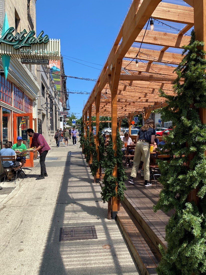 An outdoor dining area at Lost Lake Café, a restaurant in Seattle's Capitol Hill neighborhood. People can be seen enjoying their meals, along with people walking on the sidewalk in the background, on a clear, sunny day.