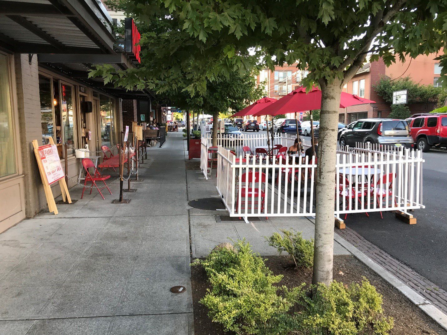 An outdoor dining area at NUE, a restaurant in Seattle's Capitol Hill neighborhood. A fenced eating area with red sun umbrellas is visible in the middle of the photo, with parked cars and buildings visible in the background.