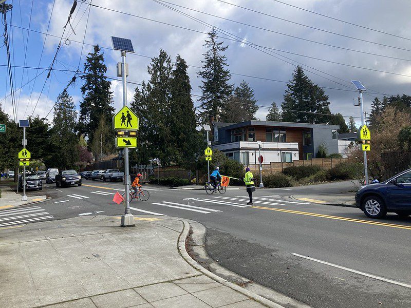 Photo of a community crossing volunteer helping stop traffic so a student and adult can safely cross the street while biking to school in Seattle.