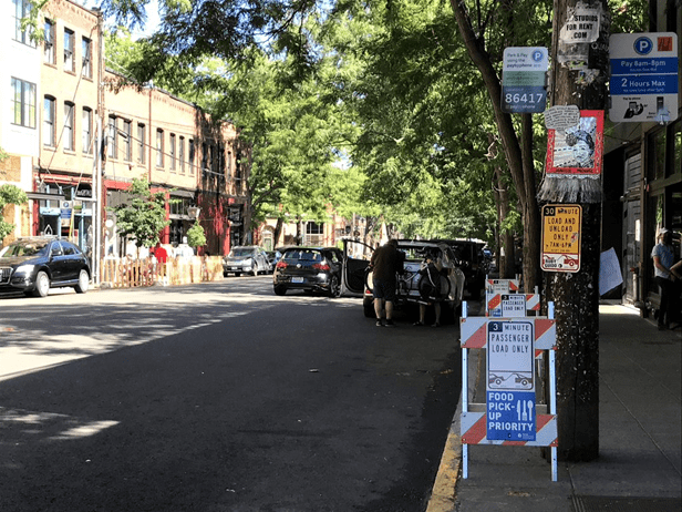 Photo of on-street paid parking and load zones in Seattle’s Ballard neighborhood. Large green trees and buildings are visible in the background, along with several cars and people in the image.