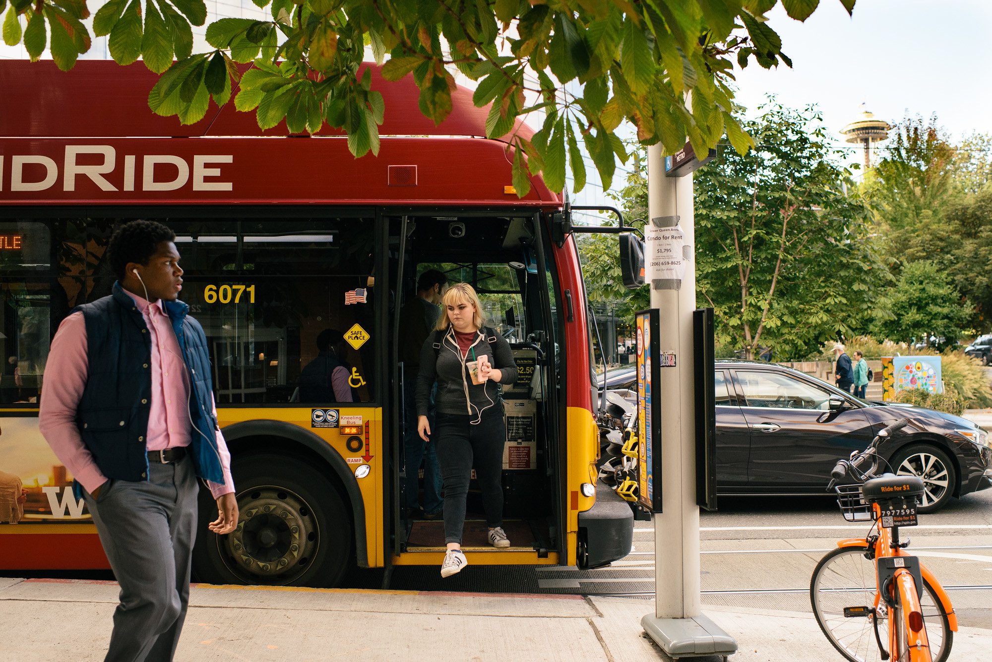 A woman steps out from a RapidRide bus in Seattle, with the Space Needle visible in the background. An orange bicycle and black car can be seen on the right-hand side of the image, with a man walking on the sidewalk in the left-hand side of the image. Green trees are visible in the background.