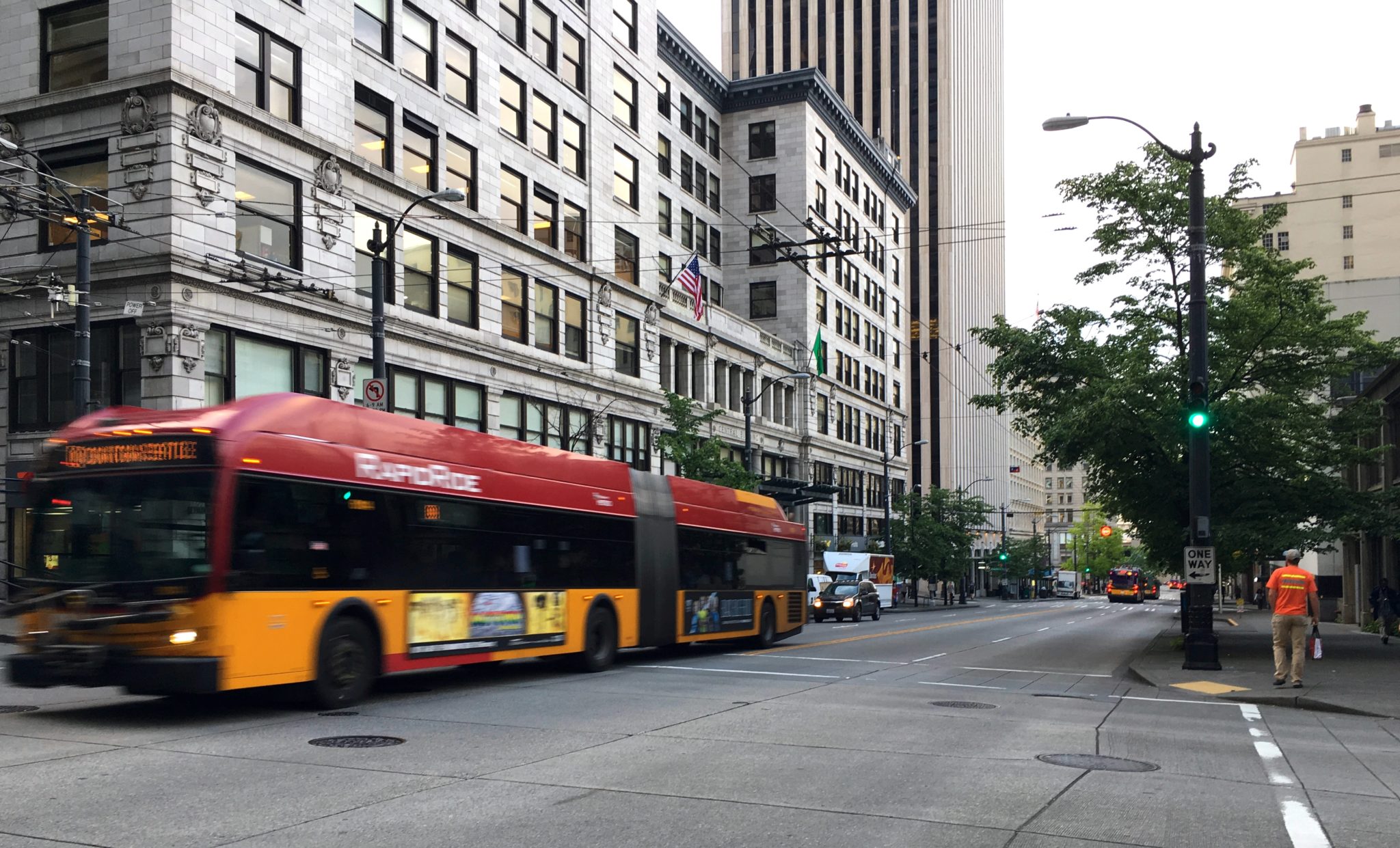 A RapidRide bus travels through downtown Seattle, along 3rd Ave. Buildings and other vehicles, as well as one person walking and wearing an orange shirt, are visible in the photo, along with several trees.