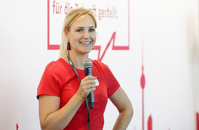 Larissa Zeichhardt speaking at an event in a red short sleeved shirt holding a microphone.
