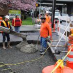 Crew members work to pave a section of the street in north Seattle. Cars can be seen to the right, driving past the work zone.
