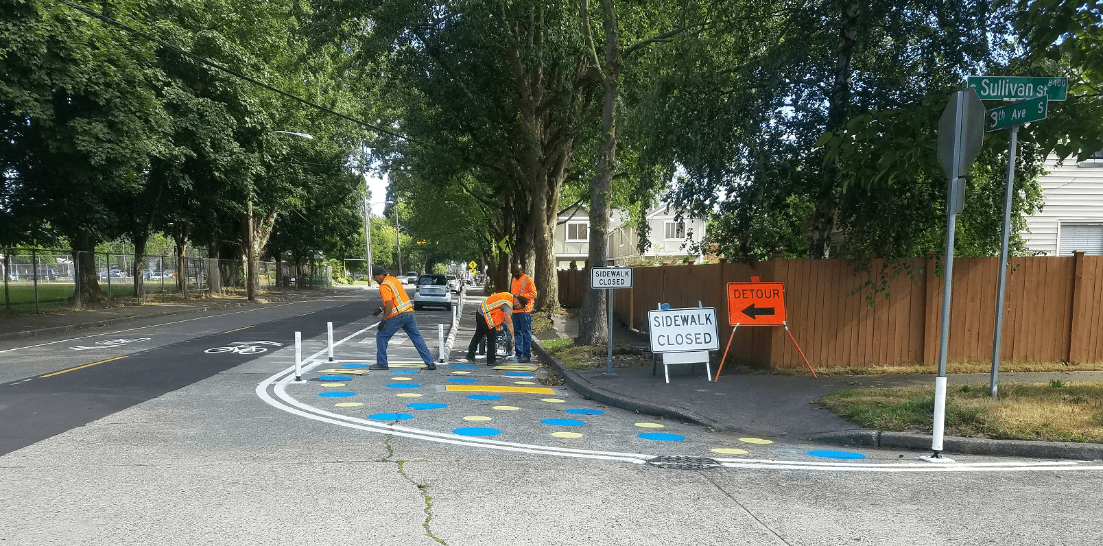 Crew members install a painted curb bulb in South Park, at 8th Ave S and S Sullivan St. Three people can be seen working, with the street visible to the left and in the foreground. Large trees are visible near the top of the image.