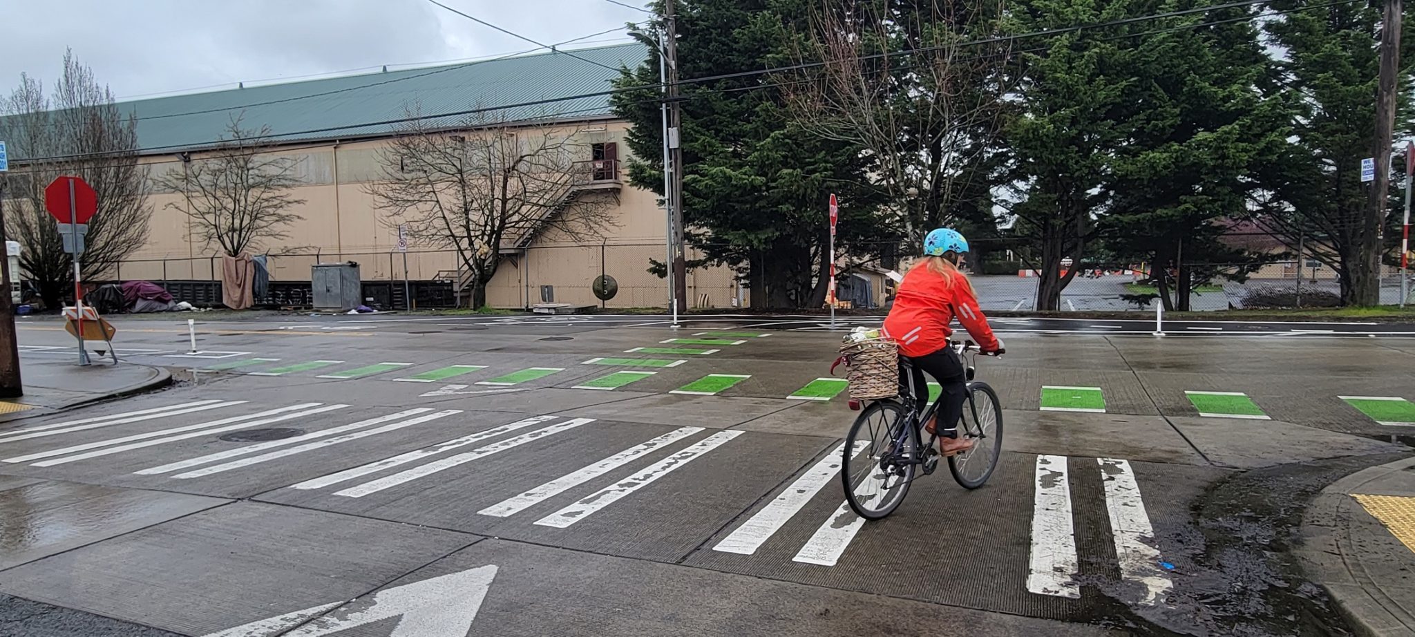 A bicyclist turns right onto Delridge Way SW. The biker is wearing a red jacket and light blue helmet. They have a small basket on the back on their bicycle. Several trees and a large building are visible in the background.