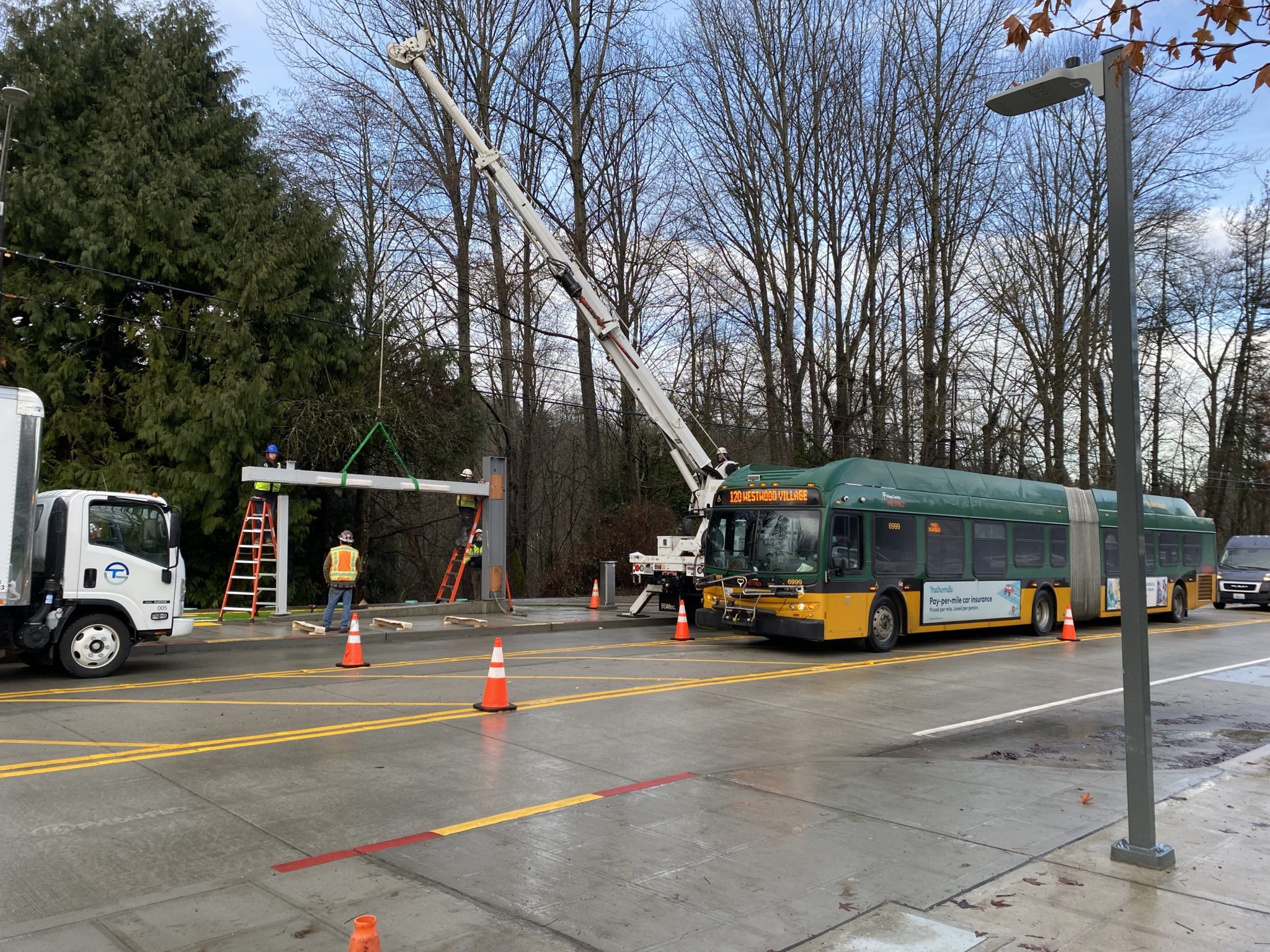 King County Metro installs a new RapidRide bus station. In the photo, several people work to install the new bus station, as traffic moves through the area in a temporary configuration, including a bus and a delivery van. Trees are visible in the background.