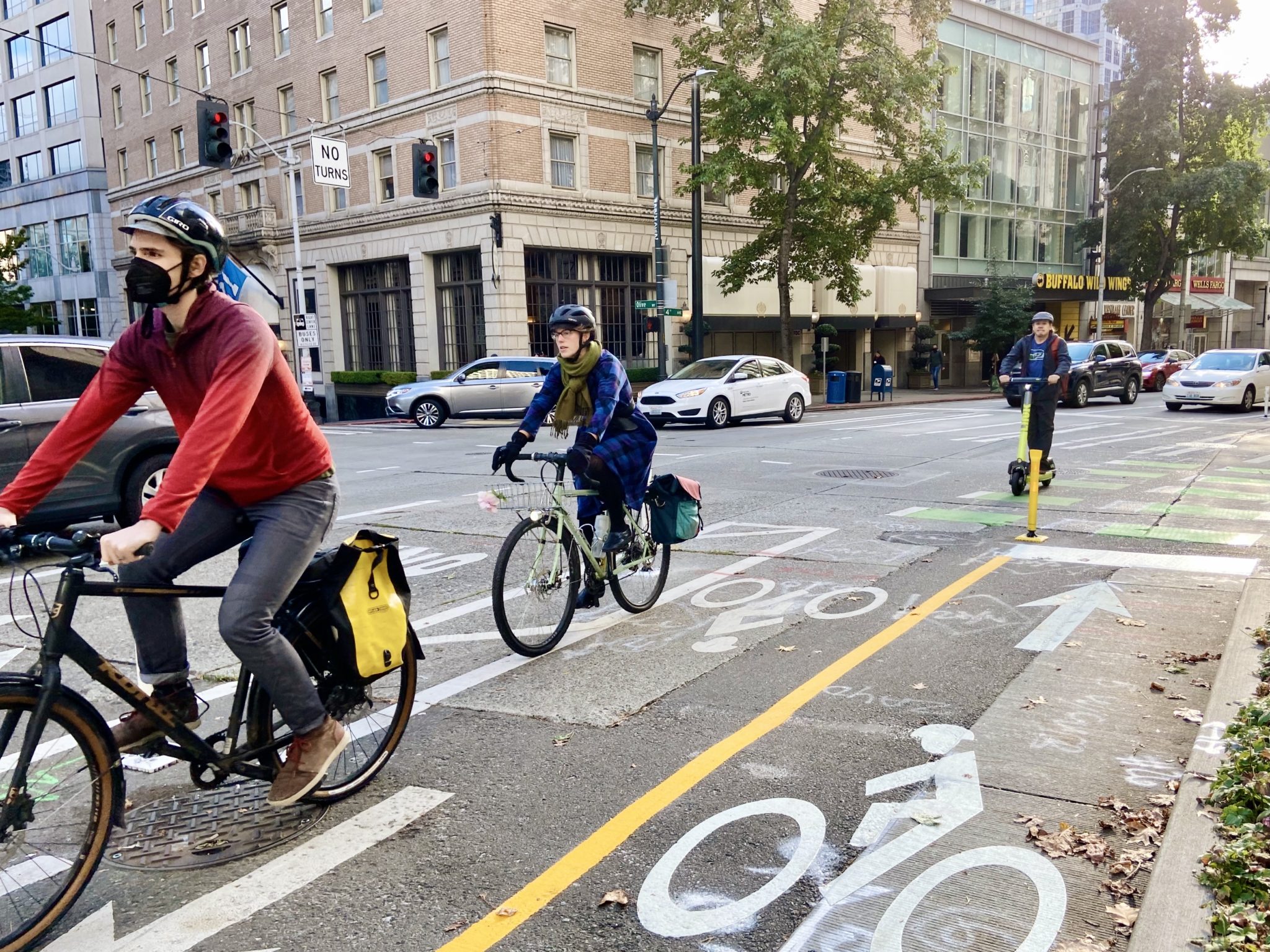 Two people ride bikes and one person rides a scooter in a protected bike lane through downtown Seattle.