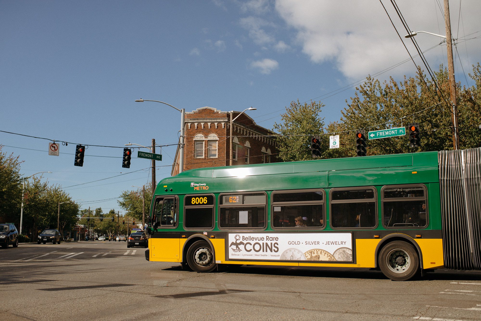 A King County Metro route 62 bus travels along Fremont Ave and Fremont Pl. Traffic signals, a large brick building, trees, and clouds, are visible in the background.