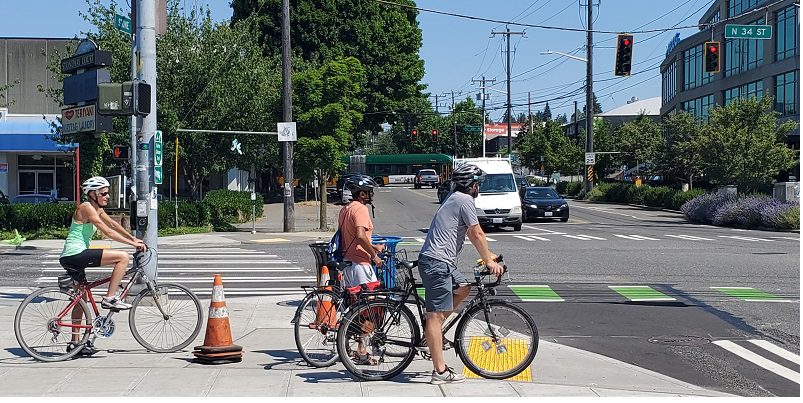 People bike along N 34th St in Fremont, on a sunny day. Cars and a bus can be seen in the background, along with large, mature trees.
