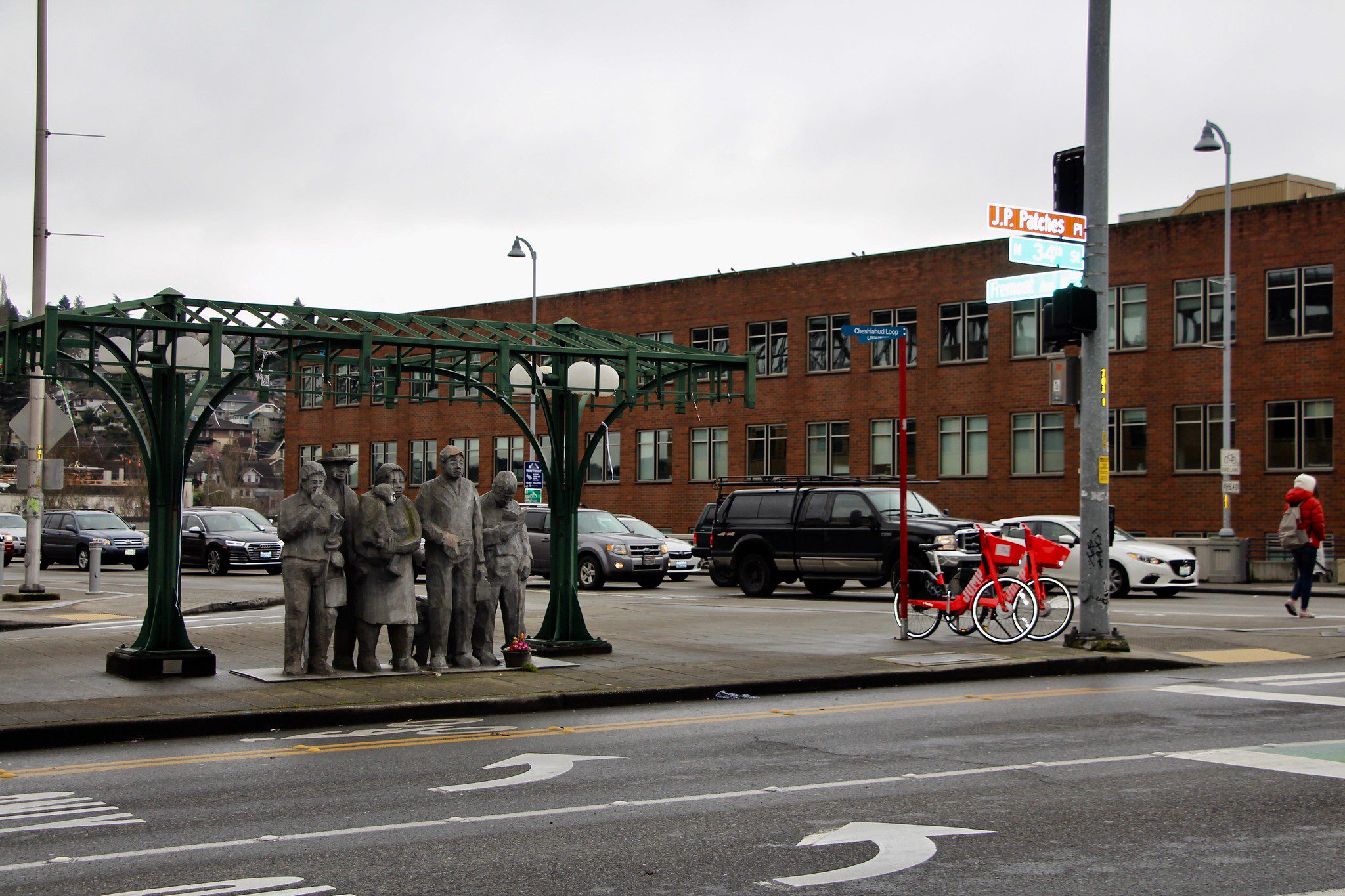The iconic Waiting for the Interurban statue in Fremont, with drivers along Fremont Ave N waiting for the light to turn green. A large brick building is located in the background, along with two red rideshare bicycles.